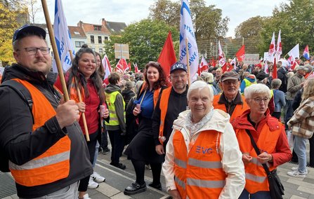 Menschenmenge auf einem Platz mit vielen roten und weißen Fahnen. Im Vordergrund stehen mehrere Personen mit orangefarbenen Warnwesten, einige halten Fahnenstangen. Im Hintergrund sind weitere Teilnehmende, Fahnen und Gebäude zu sehen.