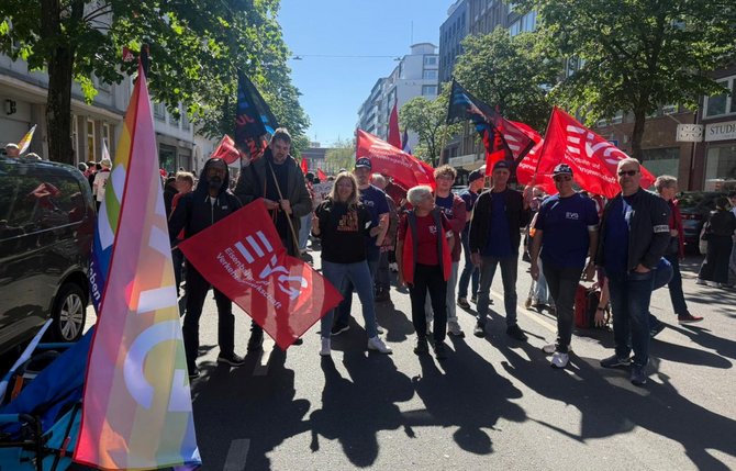 Straßenszene mit einer größeren Gruppe von Personen, die rote Fahnen mit der Aufschrift „Eisenbahn- und Verkehrsgewerkschaft“ tragen. Weitere Fahnen in unterschiedlichen Farben sind zu sehen. Die Personen stehen zwischen parkenden Autos und Gebäuden. Bäume werfen Schatten auf die Straße.