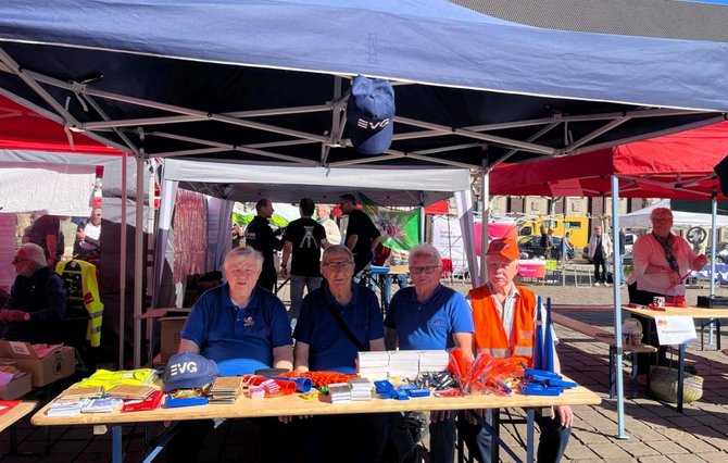 Mehrere Personen sitzen hinter einem Holztisch unter einem blauen Pavillon im Freien. Auf dem Tisch liegen Flyer, Karten, Stifte, Schlüsselanhänger, kleine Werbeartikel und Kappen mit der Aufschrift „EVG“. Über dem Tisch hängt eine blaue Kappe am Pavillongestänge. Hinter dem Tisch stehen weitere Personen, daneben sind weitere Pavillons, Fahnen und Infostände zu sehen. Der Untergrund besteht aus Pflastersteinen, im Hintergrund stehen Gebäude und weitere Teilnehmende.
