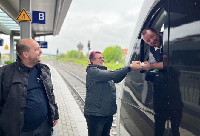  Bahnsteig an einem Bahnhof mit Gleisen im Hintergrund. Drei Personen stehen neben einem schwarzen Zug. Eine Person reicht durch ein geöffnetes Zugfenster einen kleinen Gegenstand an eine andere Person auf dem Bahnsteig. Eine weitere Person steht daneben. Am Bahnsteig sind ein Schild mit dem Buchstaben „B“ und ein Warnsymbol zu sehen.