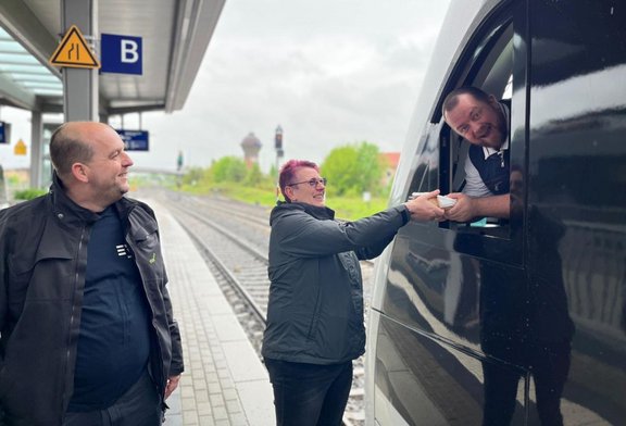  Bahnsteig an einem Bahnhof mit Gleisen im Hintergrund. Drei Personen stehen neben einem schwarzen Zug. Eine Person reicht durch ein geöffnetes Zugfenster einen kleinen Gegenstand an eine andere Person auf dem Bahnsteig. Eine weitere Person steht daneben. Am Bahnsteig sind ein Schild mit dem Buchstaben „B“ und ein Warnsymbol zu sehen.