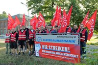 Gruppe von Personen in roten Warnwesten steht im Freien hinter einem Schild. Mehrere rote Fahnen mit „EVG“ sind hochgehalten. Auf dem Schild steht „Bahntechnikerring 90“, „GLEISBAUMECHANIK“ sowie auf einem roten Banner „WIR STREIKEN! KEIN ABSCHLUSS OHNE GEWERKSCHAFT“. Im Hintergrund sind Bäume, ein Backsteingebäude und eine Straße zu sehen.