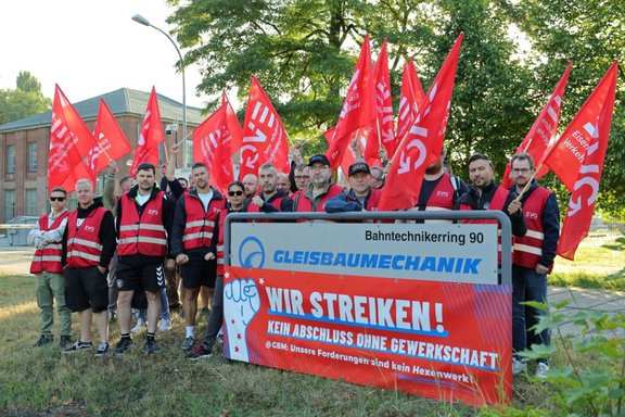 Gruppe von Personen in roten Warnwesten steht im Freien hinter einem Schild. Mehrere rote Fahnen mit „EVG“ sind hochgehalten. Auf dem Schild steht „Bahntechnikerring 90“, „GLEISBAUMECHANIK“ sowie auf einem roten Banner „WIR STREIKEN! KEIN ABSCHLUSS OHNE GEWERKSCHAFT“. Im Hintergrund sind Bäume, ein Backsteingebäude und eine Straße zu sehen.