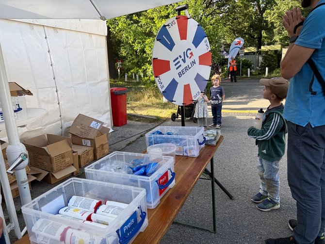 Ein Kind steht vor einem Glücksrad an einem Tisch mit Preisen, während eine weitere Person daneben steht. Im Vordergrund steht ein Kind vor einem großen, runden Glücksrad, das auf einem Ständer befestigt ist. Auf dem Glücksrad ist gut lesbar der Schriftzug „EVG Berlin“ sowie farbige Felder in Blau, Weiß und Rot. Rechts daneben steht eine weitere Person, die das Kind beobachtet. Vor dem Glücksrad befindet sich ein langer Holztisch, auf dem mehrere durchsichtige Plastikboxen stehen. In den Boxen liegen verschiedene Gegenstände, zum Beispiel Becher, kleine verpackte Artikel und andere Preise. Einige Boxen sind mit dem Wort „Preis“ beschriftet. Links im Bild ist ein weißes Zelt aufgebaut. Am Boden neben dem Zelt liegen Kartons und Verpackungen. Im Hintergrund stehen weitere Menschen, darunter auch ein weiteres Kind. Außerdem sind Bäume, eine Straße und eine weitere Fahne mit ähnlichem Aufdruck wie das Glücksrad zu sehen.