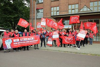 Gruppe von Demonstrierenden vor einem Backsteingebäude. Viele rote EVG-Fahnen werden hochgehalten. Im Vordergrund ein großes rotes Banner mit der Aufschrift „WIR STREIKEN! KEIN ABSCHLUSS OHNE GEWERKSCHAFT“. Mehrere Schilder mit der Überschrift „MERKT EUCH DAS:“ sind sichtbar.