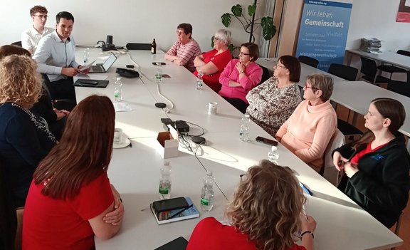 Mehrere Personen sitzen um einen großen Konferenztisch in einem hellen Raum. Auf dem Tisch stehen Wasserflaschen, Smartphones, eine Laptop-Station sowie Kabel. Eine Person am Kopfende des Tisches hält ein Tablet oder Dokument und spricht zu der Gruppe. Im Hintergrund stehen Stühle, ein Regal mit Zeitschriftenstapeln und ein Banner mit lesbarem Text: „Wir leben Gemeinschaft“.