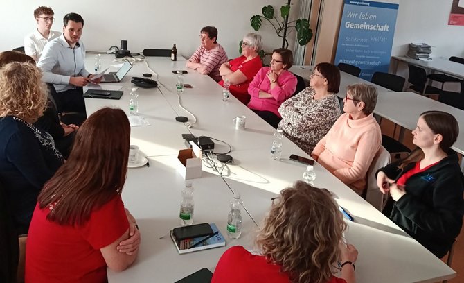 Mehrere Personen sitzen um einen großen Konferenztisch in einem hellen Raum. Auf dem Tisch stehen Wasserflaschen, Smartphones, eine Laptop-Station sowie Kabel. Eine Person am Kopfende des Tisches hält ein Tablet oder Dokument und spricht zu der Gruppe. Im Hintergrund stehen Stühle, ein Regal mit Zeitschriftenstapeln und ein Banner mit lesbarem Text: „Wir leben Gemeinschaft“.