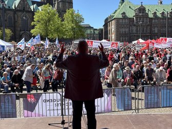 Blick von einer Bühne auf einen großen Platz mit vielen sitzenden und stehenden Menschen. Im Vordergrund steht eine Person mit dem Rücken zur Kamera am Rednerpult, beide Arme sind erhoben. Vor der Bühne sind Absperrgitter angebracht. In der Menschenmenge sind zahlreiche Fahnen und Banner zu sehen, unter anderem mit Aufschriften wie „IG Metall“ und weiteren Gewerkschaftslogos. Im Hintergrund stehen historische Gebäude mit grünen Dächern, eine Kirche und Bäume unter blauem Himmel.