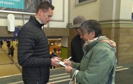 In einer Bahnhofshalle stehen zwei Personen einander gegenüber. Eine Person hält ein Klemmbrett und schreibt mit einem Stift, die andere hält ein Blatt Papier. Beide Gesichter sind verpixelt. Im Hintergrund sind Treppenabgänge, Durchgänge und weitere Personen zu sehen. An der Wand hängt ein Plakat mit teilweise lesbarem Text.