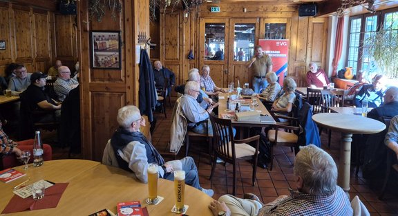 Großer Gastraum mit Holzvertäfelung, Holzbalken und Holzmöbeln. Mehrere Personen sitzen an runden und rechteckigen Tischen, auf den Tischen stehen Biergläser, Wasserflaschen, Gläser und Papierunterlagen. Eine Person steht neben einem Tisch und hält ein Mikrofon oder ein ähnliches Gerät. Im Hintergrund sind Fenster, eine Glastür und weitere sitzende Personen zu sehen. An der rechten Seite steht ein aufgerolltes Banner mit dem sichtbaren Text „EVG“ und „Bezirksverband Niederrhein“. Deckenlampen, Wanddekorationen und Pflanzen sind im Raum verteilt.