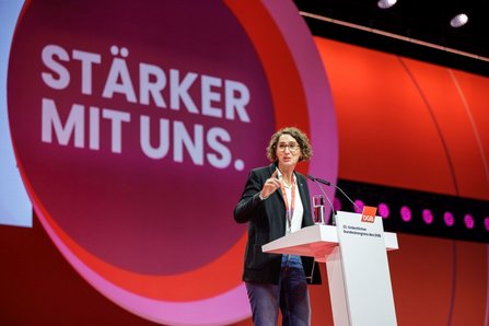 erson steht an einem weißen Rednerpult auf einer Bühne. Auf dem Pult sind zwei Mikrofone, ein Glas Wasser und ein rotes Logo mit der Aufschrift „DGB“. Im Hintergrund dominiert ein großer roter Kreis mit weißem Text „STÄRKER MIT UNS.“ Die Bühne ist in Rot‑ und Orangetönen beleuchtet, mit sichtbaren Scheinwerfern im oberen Bereich. Die Person trägt eine dunkle Jacke, ein helles Oberteil und Jeans und hebt eine Hand mit ausgestrecktem Zeigefinger.