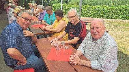 Mehrere Personen sitzen an langen Holztischen unter einer überdachten Terrasse. Auf den Tischen stehen umgedrehte Gläser und rote Platzsets. Im Vordergrund sitzen drei Personen an einem Tisch, im Hintergrund verläuft eine Hecke entlang des Geländes.