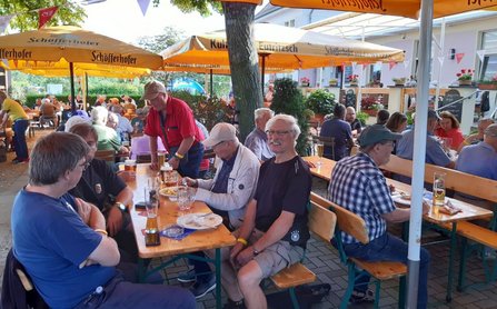 Außenbereich mit mehreren langen Holztischen und Bänken unter gelben Sonnenschirmen. Viele Personen sitzen oder stehen zwischen den Tischen. Auf den Tischen stehen Gläser, Teller und Getränke. In der Mitte verläuft ein Baumstamm zwischen den Sitzbereichen. Auf den Sonnenschirmen ist der Schriftzug „Schöfferhofer“ sichtbar.