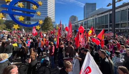 Dicht gedrängte Menschenmenge vor dem großen blauen Euro-Symbol und modernen Bürogebäuden. Viele rote Fahnen und rote Ballons ragen über die Menge hinaus. Im Vordergrund halten mehrere Personen rote und weiße Fahnen, darunter eine weiße Fahne mit rotem ver.di-Logo. Die Szene ist von Sonnenlicht geprägt.